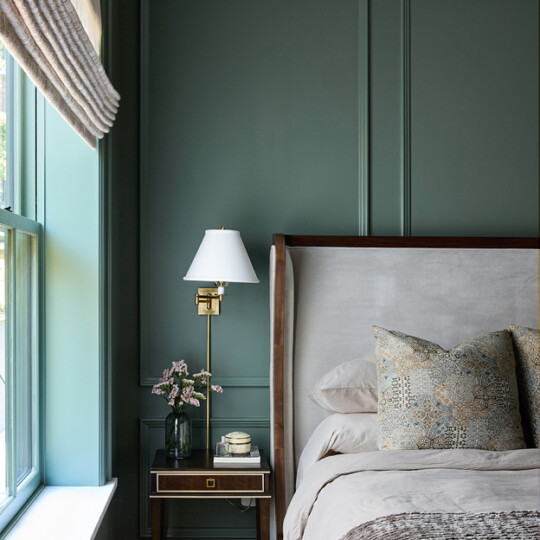 Bedroom with green paneled walls, beige upholstered headboard, decorative pillow, brass wall lamp, nightstand with flowers, near a sunny window.