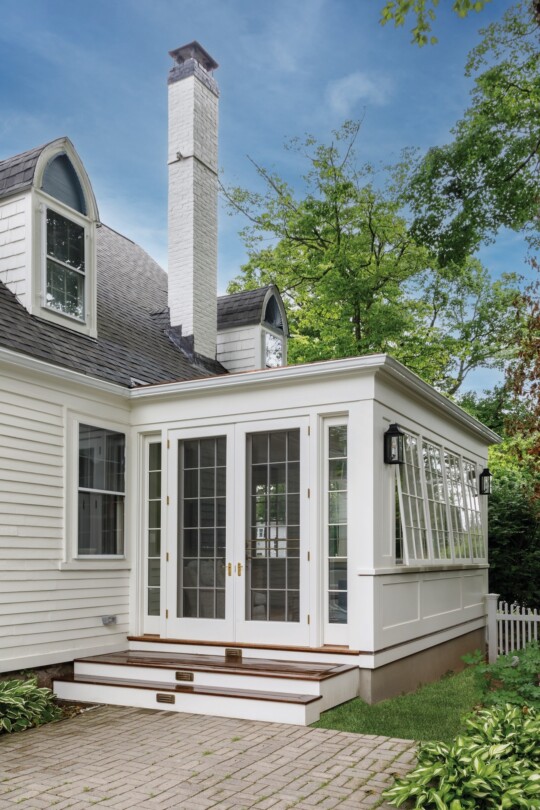 White house extension with large glass French doors, a brick chimney, and dormer windows; surrounded by green trees, a paved patio, and a white picket fence.