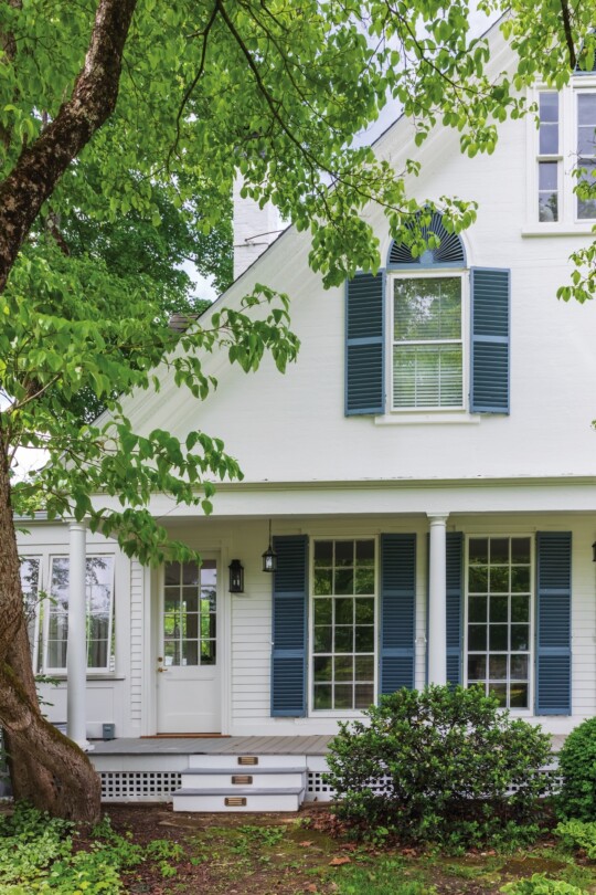 White two-story house with blue shutters, large center window, covered porch, and green landscaping; prominent tree branches frame the facade.