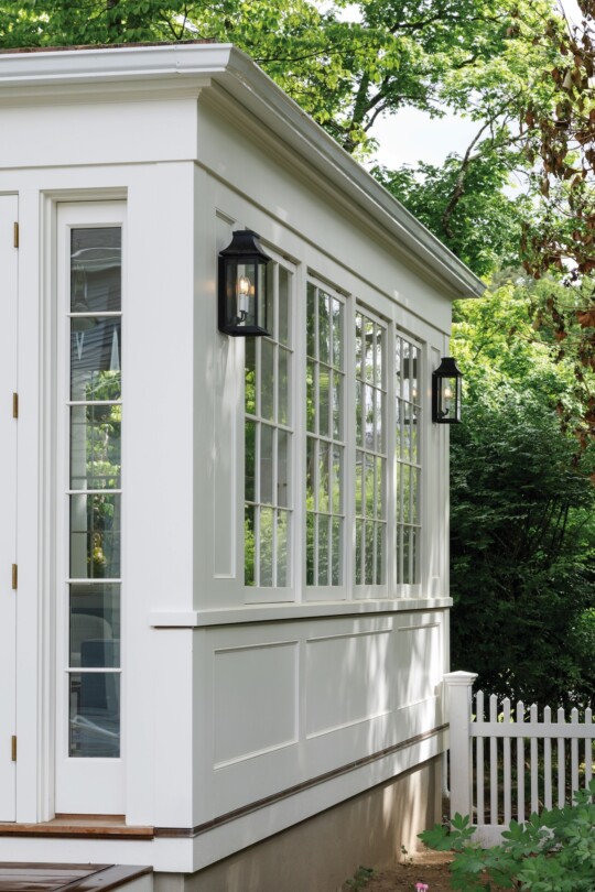 White framed sunroom with large multi-pane windows, black wall lanterns, and a white picket fence; lush green trees and shrubs surround the bright exterior.