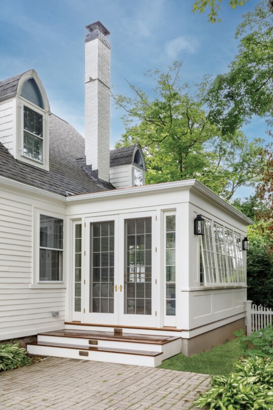 White residential home addition with extensive glass doors and windows, tall white chimney, dark roof with dormers, brick patio, and surrounding green trees under clear blue sky