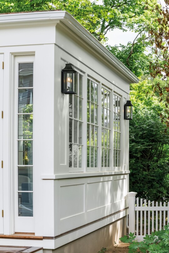 Modern white garden shed with large multi-pane windows and black lantern sconces, surrounded by lush green trees and a white picket fence.