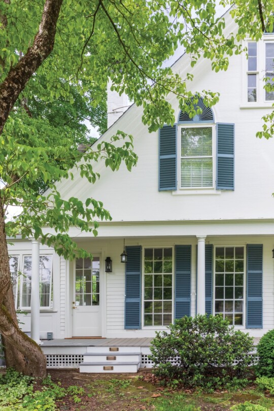 White farmhouse exterior with blue-green shutters, covered porch with white columns, multiple windows, and a large tree with green foliage in the foreground