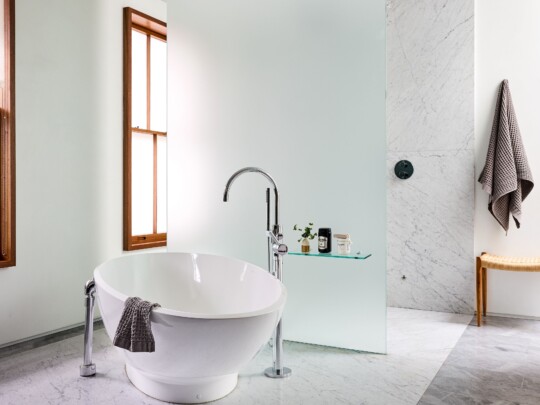 Modern bathroom with a freestanding white tub, curved chrome faucet, marble floor, frosted glass partition, a wooden-framed window, and a towel on the tub.