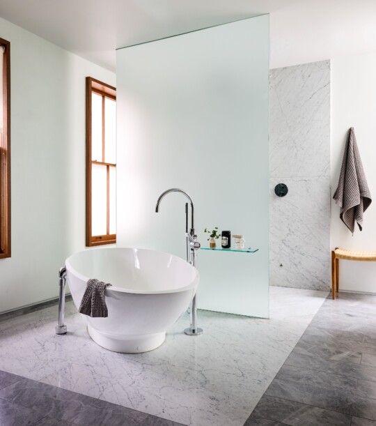 Modern bathroom with a freestanding white tub, curved chrome faucet, marble floor, frosted glass partition, a wooden-framed window, and a towel on the tub.
