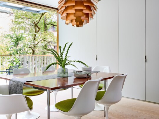 Bright dining room with a wooden table, tulip chairs with green cushions, a pendant lamp, and a vase of greenery; windows overlook trees.
