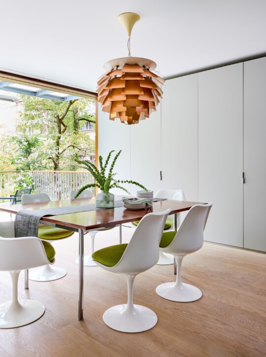 Bright dining room with a wooden table, tulip chairs with green cushions, a pendant lamp, and a vase of greenery; windows overlook trees.
