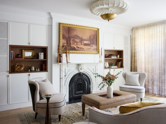 Bright living room with white built-in cabinets around a decorative fireplace, a framed landscape painting, beige armchairs, a brown leather coffee table, and sheer curtains.