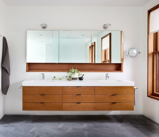 Modern bathroom with white double-sink vanity, warm wood drawers, a wide mirrored cabinet, potted plant, dark tile floor, and a wooden-framed window.