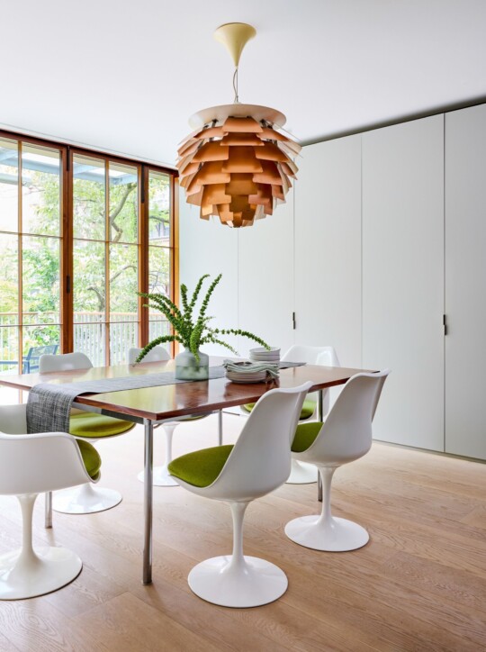 Bright dining room with a wooden table and white tulip chairs with green cushions, a layered wood pendant light, large windows, and white cabinets.