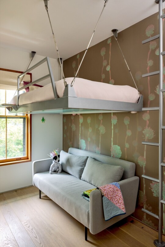 Room with a metal loft bed suspended from the ceiling over a gray sofa, a colorful quilt on the arm, a crown-topped stuffed animal, and tree wallpaper.