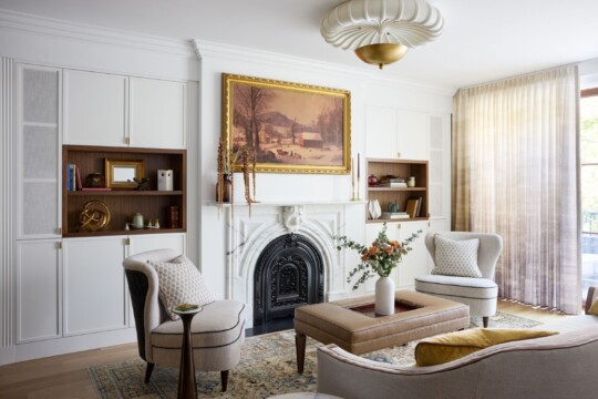 Bright living room with white built-ins, a gold-framed landscape painting above a decorative fireplace, two cream chairs, and a tan coffee table.