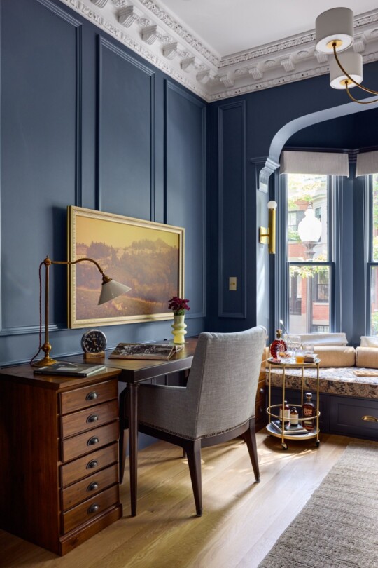 Blue-paneled room with ornate crown molding, a wooden desk, gray upholstered chair, brass lamp, landscape painting, and a window seat with cushions beside a bar cart.