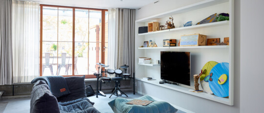 Bright living room with a large window and sheer curtains, a blue cushioned lounge chair, a drum kit by the window, and a white built-in shelf with a TV.