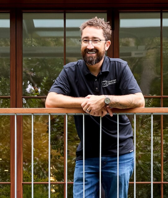 Person with short hair and beard, wearing a dark polo and glasses, leaning on a railing outside a glass-walled building and smiling.