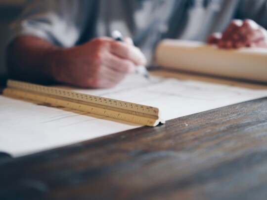 Close-up of a person writing on a large sheet of paper, with a wooden ruler laid across and a rolled blueprint beside them on a wooden desk.