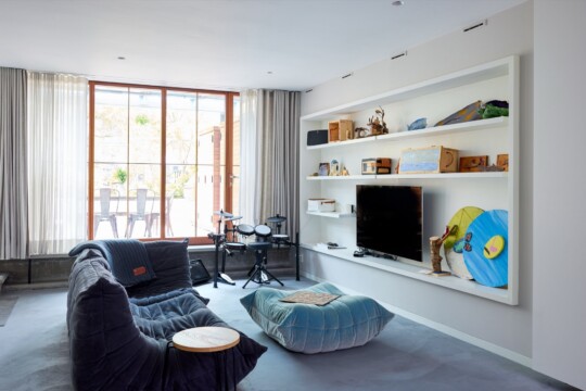 Bright modern living room with a large window, sheer curtains, white built-in shelves and TV, a dark blue cushioned sofa, a blue bean bag, and a small drum set by the window.