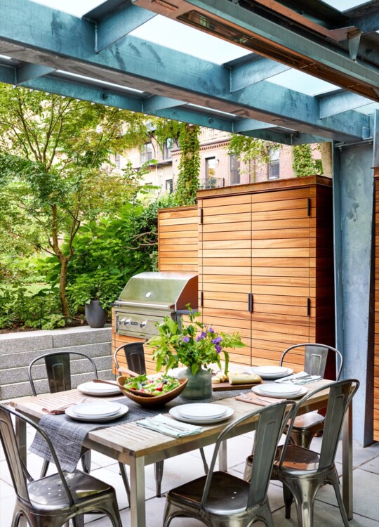 Outdoor dining area under a blue steel frame with a wooden cabinet and grill; table set with plates, cutlery, a salad bowl, and a vase of flowers.