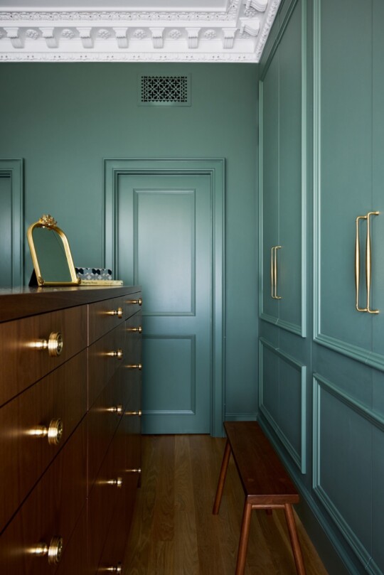 Teal paneled dressing room with a dark wood dresser featuring brass knobs, a gold-framed mirror, a wooden bench, and ornate white ceiling molding.
