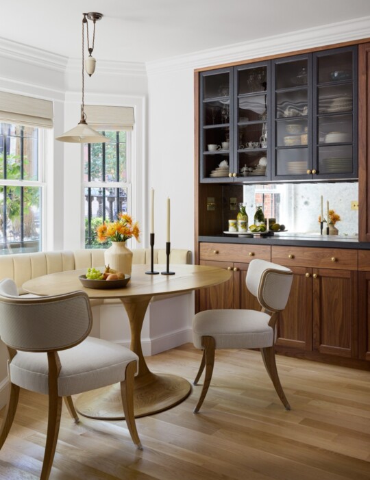Bright dining nook with a round wooden table, cream upholstered chairs, a beige banquette, and dark glass-front cabinets; sun streams through large windows.