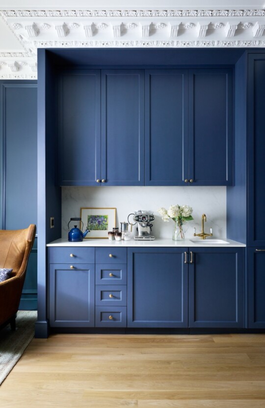 Blue kitchen cabinets with brass hardware and marble counter; a coffee machine, kettle, a vase of flowers, framed art, and a gold faucet.
