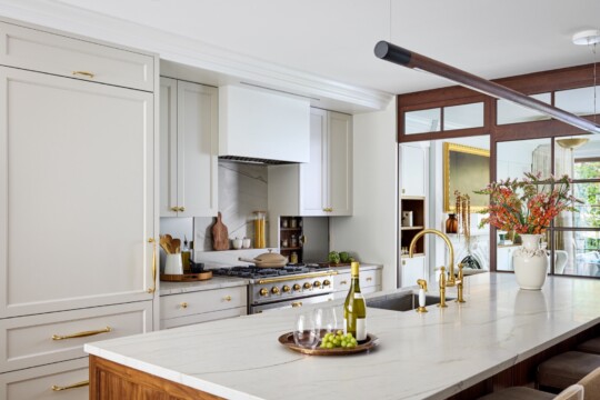 Bright kitchen with white cabinets, marble island, and gold hardware; brass faucet, a wine bottle on a tray with grapes, and a vase of flowers.