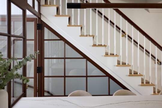Diagonal white staircase with wooden handrail and white spindles, glass-paneled wall on left, marble counter in foreground, and eucalyptus plant.