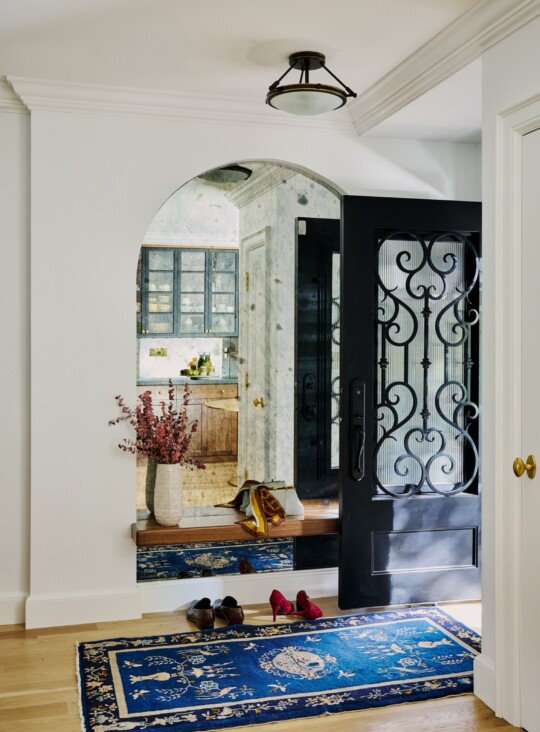 Bright entryway with a rounded arch opening to a kitchen; black decorative iron door, a vase of dried flowers, and a blue patterned rug with shoes.