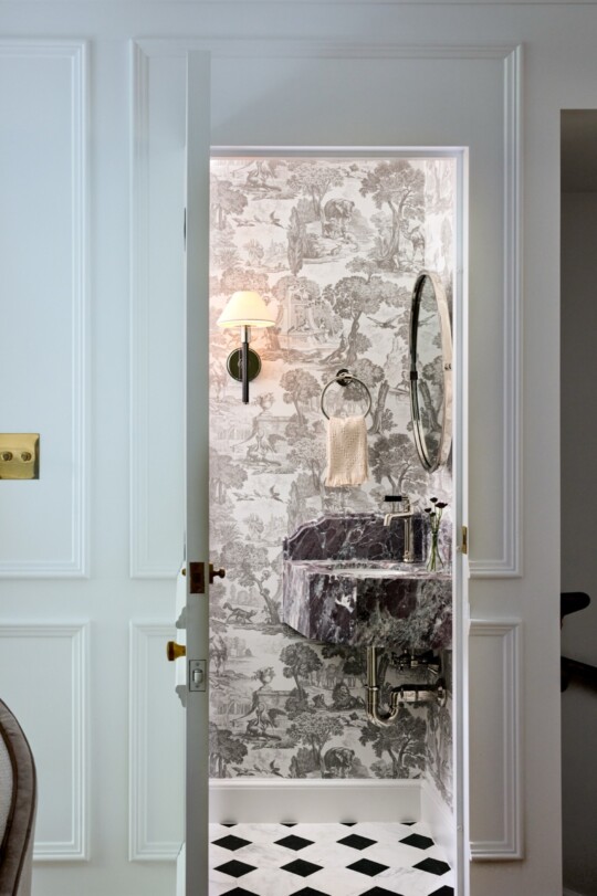 Entering a small bathroom with a black marble wall-mounted sink, round mirror, wall lamp, towel ring with a towel, toile wallpaper, and a black‑and‑white checkered floor.