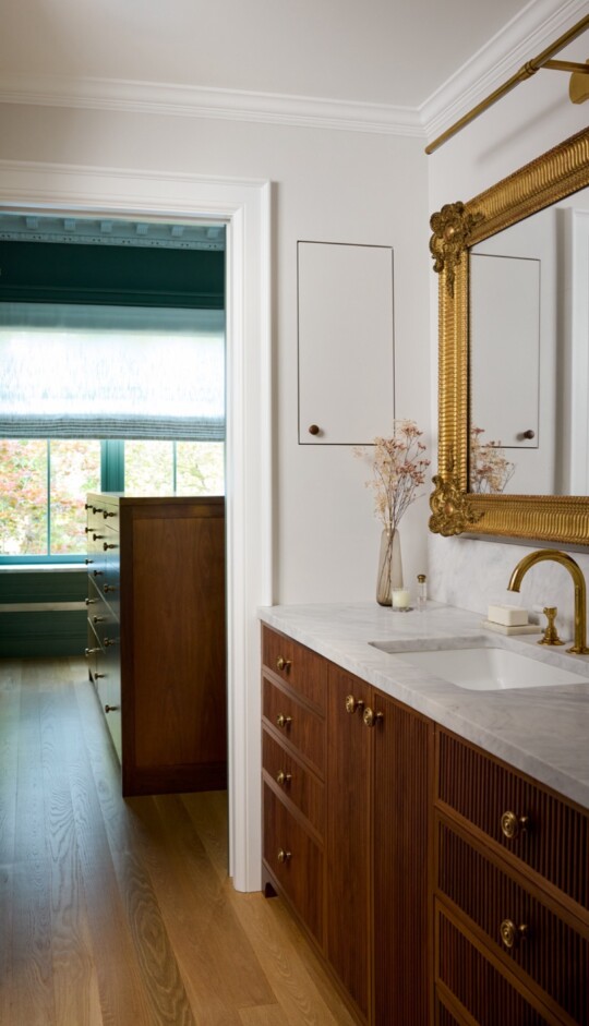 Elegant bathroom with dark wood vanity, white marble countertop, and a large ornate gold-framed mirror; dried flowers in a vase by the gold faucet.