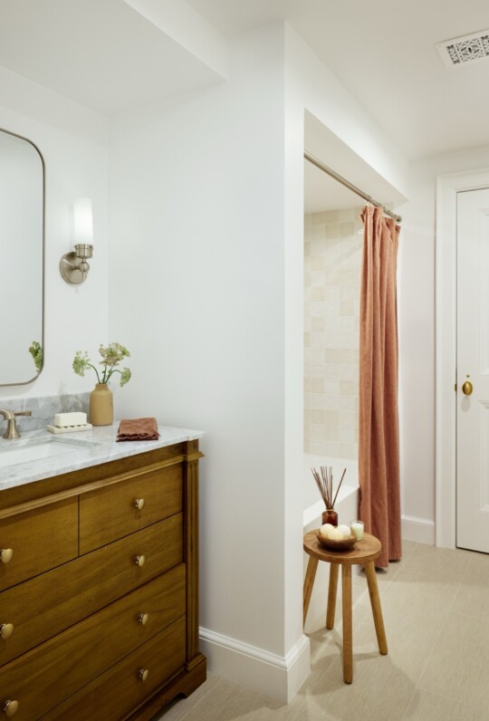 Bright bathroom with a wooden vanity, marble countertop, oval mirror, and a small plant; folded towel, pink curtain shower, and a round stool.