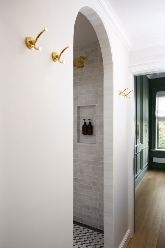 Arched shower alcove with gray subway tiles, built-in niche holding brown bottles, and a gold showerhead; white walls, wooden floor, and dark green cabinetry nearby.