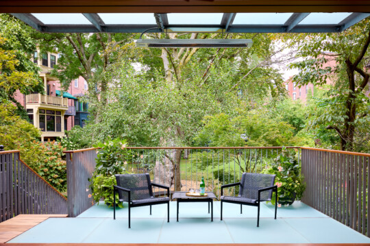 Balcony with two black chairs and a small table, bottle on the table, surrounded by green trees and neighboring apartment buildings.